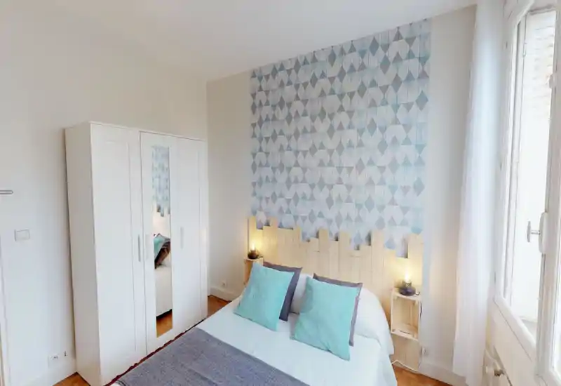 Bright, cozy bedroom with patterned accent wall, wooden headboard and double bed dressed in white linens and aqua cushions. Large window provides abundant natural light and a clean, airy atmosphere—ideal for rental photos.