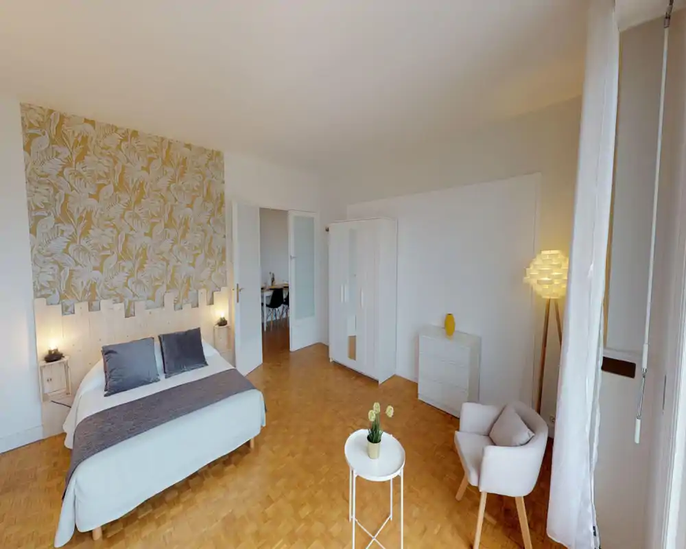 Chambre spacieuse et lumineuse avec grand lit double, mur à motif et éclairage d'appoint chaleureux. Parquet propre, armoire et fauteuil confortable pour une ambiance moderne et apaisante.