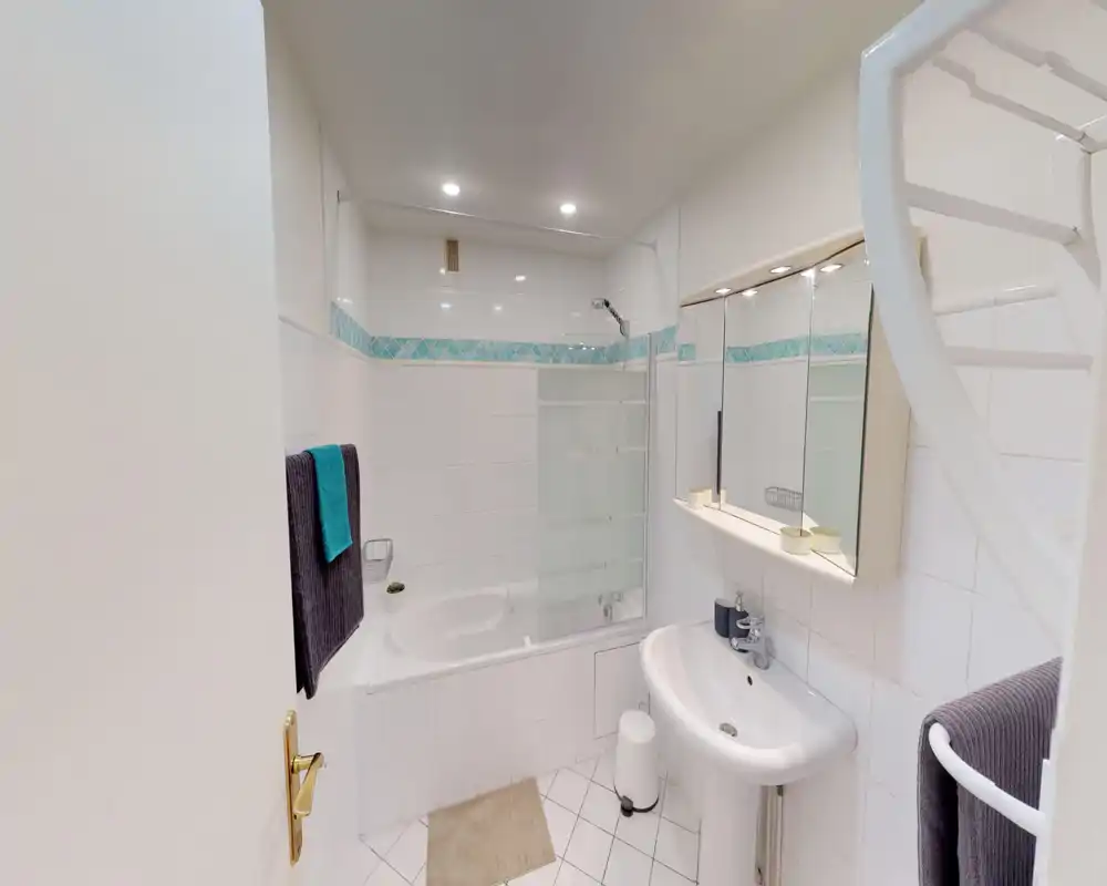 Bagno compatto e pulito con vasca-doccia, lavabo a colonna e specchiera con mobiletto. Piastrelle bianche e fascia decorativa turchese donano luminosità e freschezza.