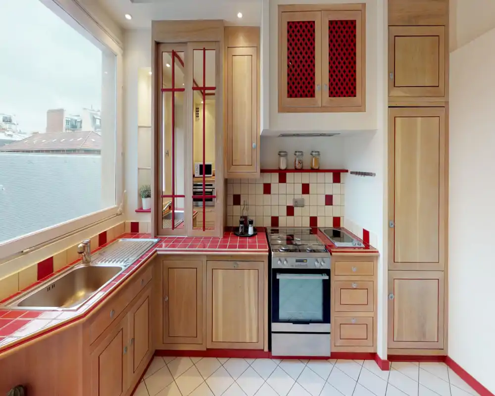 Cucina luminosa e compatta con pensili in legno, piano in piastrelle rosse e forno in acciaio inox. Ampia finestra che offre luce naturale e vista sulla città.