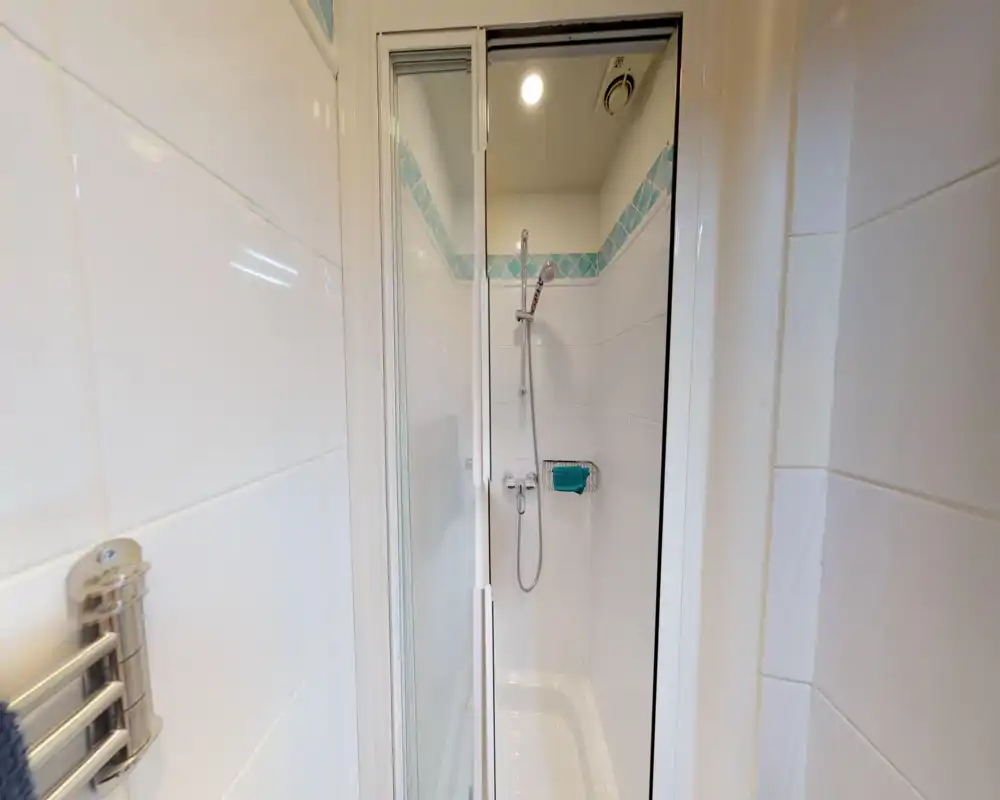 Doccia piastrellata compatta con porta scorrevole in vetro, soffione regolabile e piccolo portasapone a muro; piastrelle bianche pulite con fascia decorativa blu.