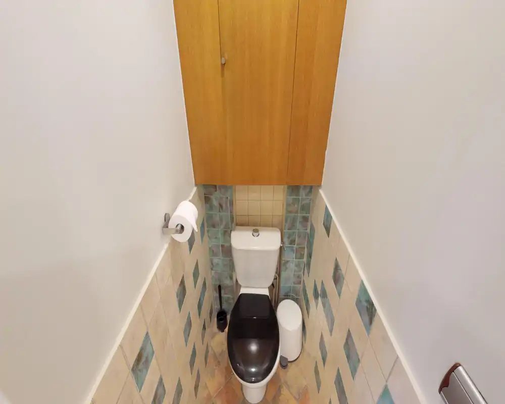 Bagno compatto e ben illuminato con vista ravvicinata sul WC, piastrelle fino a metà parete e un armadietto in legno sopra.