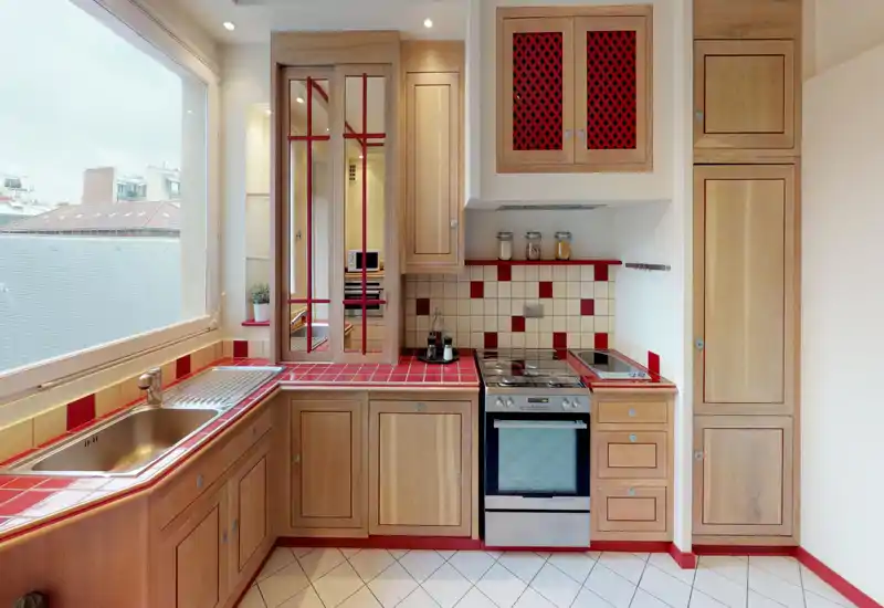 Cucina luminosa e compatta con pensili in legno, piano in piastrelle rosse e forno in acciaio inox. Ampia finestra che offre luce naturale e vista sulla città.