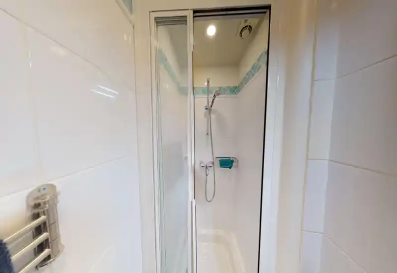 Doccia piastrellata compatta con porta scorrevole in vetro, soffione regolabile e piccolo portasapone a muro; piastrelle bianche pulite con fascia decorativa blu.