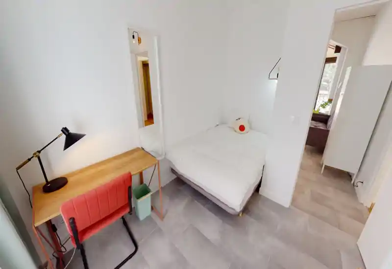 Camera da letto luminosa e compatta con un comodo letto matrimoniale, lenzuola bianche e una piccola scrivania in legno accanto a una sedia corallo — ideale per una singola persona o una coppia. L'ambiente è arioso e pulito, con illuminazione moderna e toni neutri.
