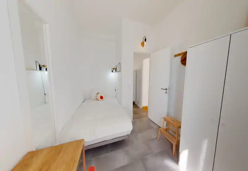 Camera da letto singola luminosa e minimal con letto ben rifatto, applique da lettura a parete, specchio a figura intera e armadio compatto — ideale per un soggiorno accogliente da solo.