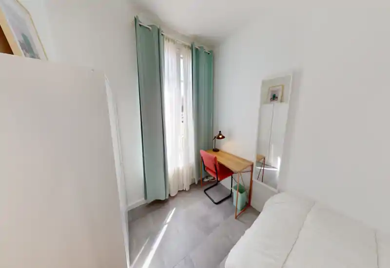Angolo di camera da letto luminoso con vista parziale del letto, una scrivania in legno e una sedia rossa davanti a una finestra alta con tende leggere e color menta — stile minimal e ottima luce naturale.