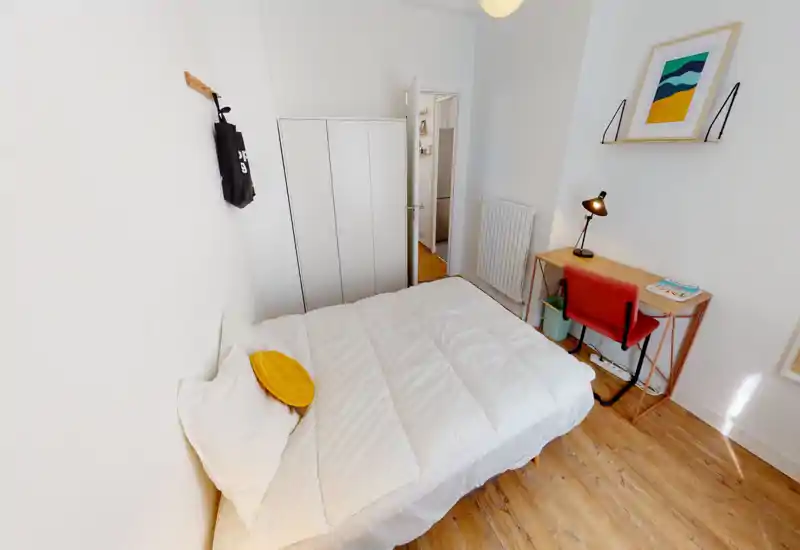 Camera da letto luminosa e minimale con letto doppio rifatto, pavimento in legno e una scrivania piccola vicino al muro — ideale per una persona o una coppia che cerca uno spazio compatto e ben illuminato.