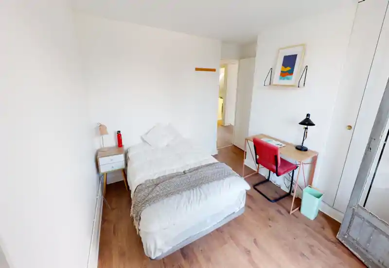 Camera da letto luminosa e minimal con letto matrimoniale al centro, comodino e piccolo angolo studio. Pareti bianche, pavimento in legno e tessili semplici per un’atmosfera moderna e rilassante.