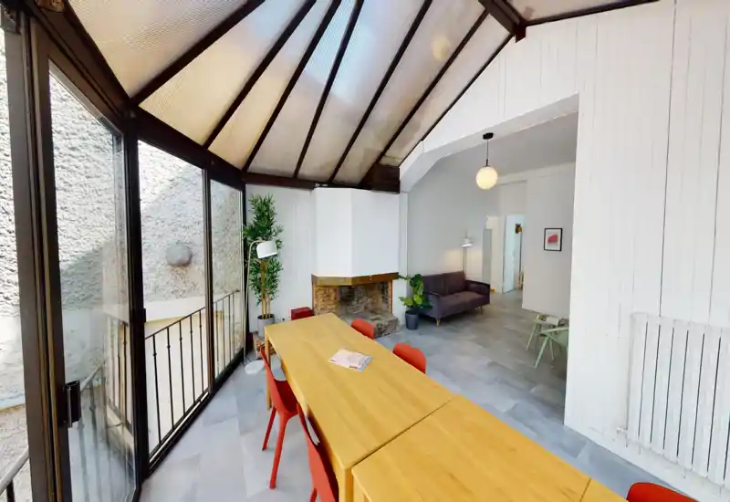 Ampio e luminoso soggiorno/pranzo con un lungo tavolo in legno e sedie rosse sotto una struttura vetrata. L'ambiente ha un divano accogliente, piante e un camino decorativo, perfetto per pranzi e relax.