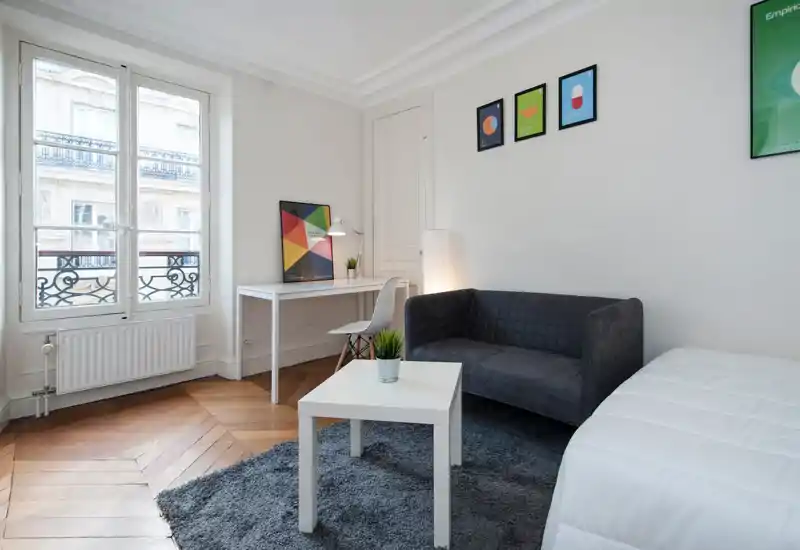 Bright, minimal bedroom with a white desk, gray sofa and a neatly made bed at the right edge; large window and parquet floor create an airy, modern feel.