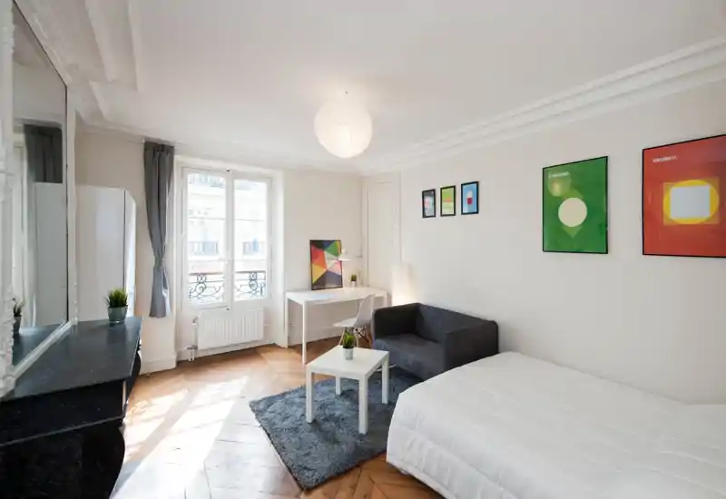 Bright, minimal bedroom with a comfortable double bed in the foreground, a small sofa and a desk by the window. Natural light, parquet flooring and framed artwork create a modern, airy studio feel ideal for short-term rentals.