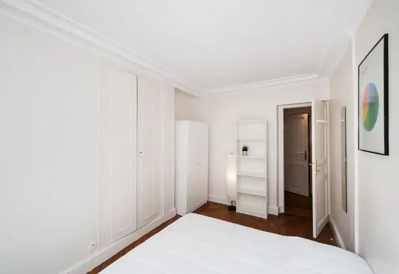Bright, minimalist bedroom with a white bed in the foreground, built-in wardrobe, freestanding wardrobe and shelving, and warm parquet flooring.