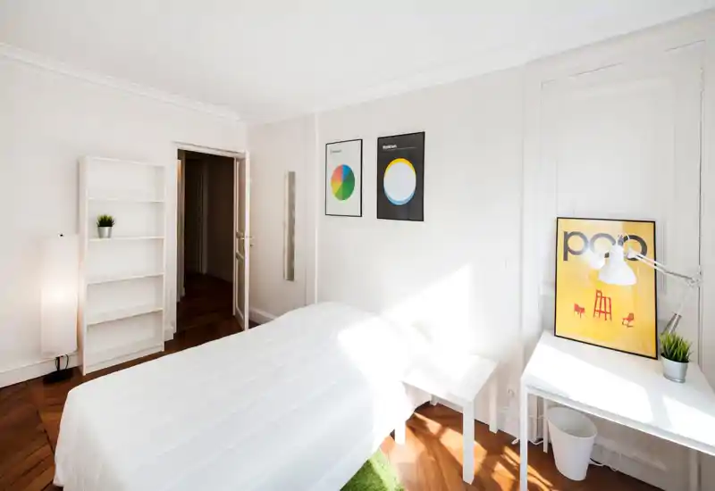 Bright, minimalist bedroom with a neatly made white bed, wooden floor and simple modern decor including framed prints and a small desk by the window.