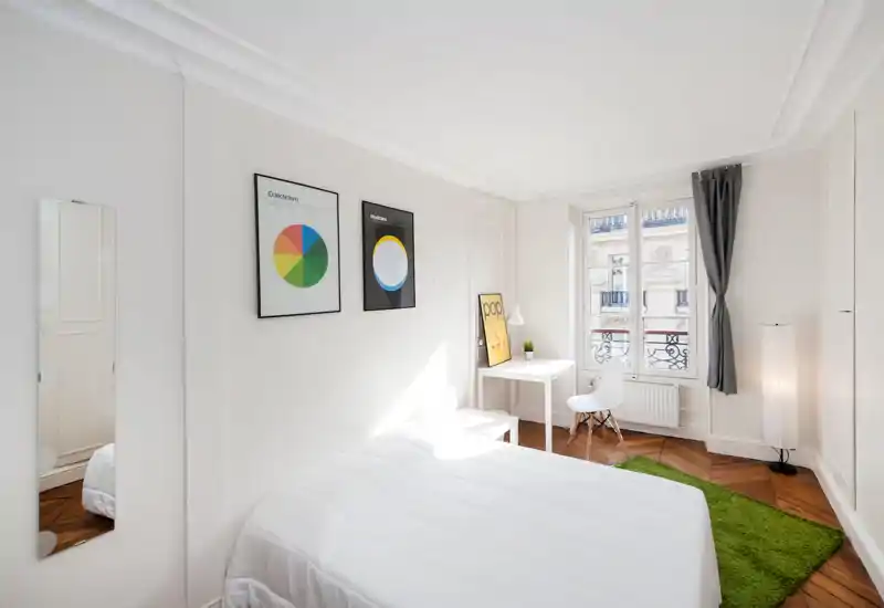 Bright and airy bedroom with a clean white bed, a small desk by the window, and warm wood floors. Large window provides plenty of daylight; minimalist decor and a green rug add a cozy touch.