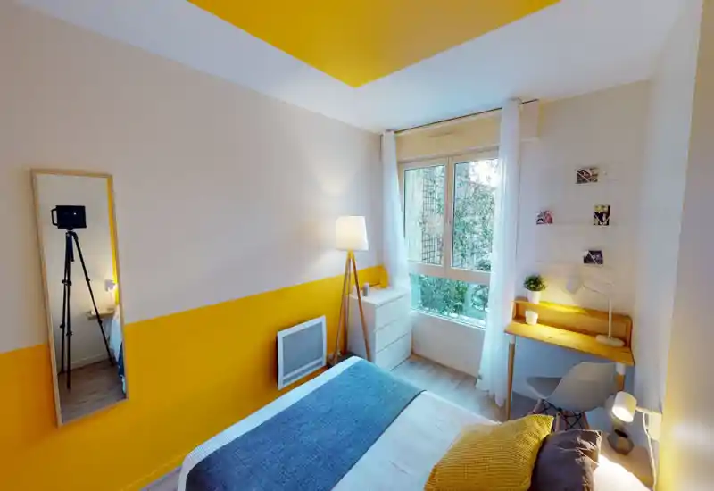 Camera da letto luminosa e curata con letto matrimoniale, lampade da comodino, una piccola scrivania vicino alla finestra e pareti accentate gialle che creano un ambiente accogliente e moderno.