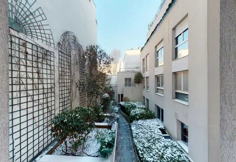 Corte interna soleggiata con vialetto stretto, siepi potate e una leggera neve nei aiuole — un'area esterna tranquilla tra gli edifici.
