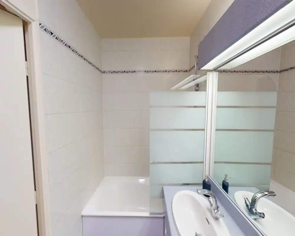 Bagno luminoso e pulito con vasca e parete doccia in vetro, lavabo con specchio e rubinetteria cromata moderna.