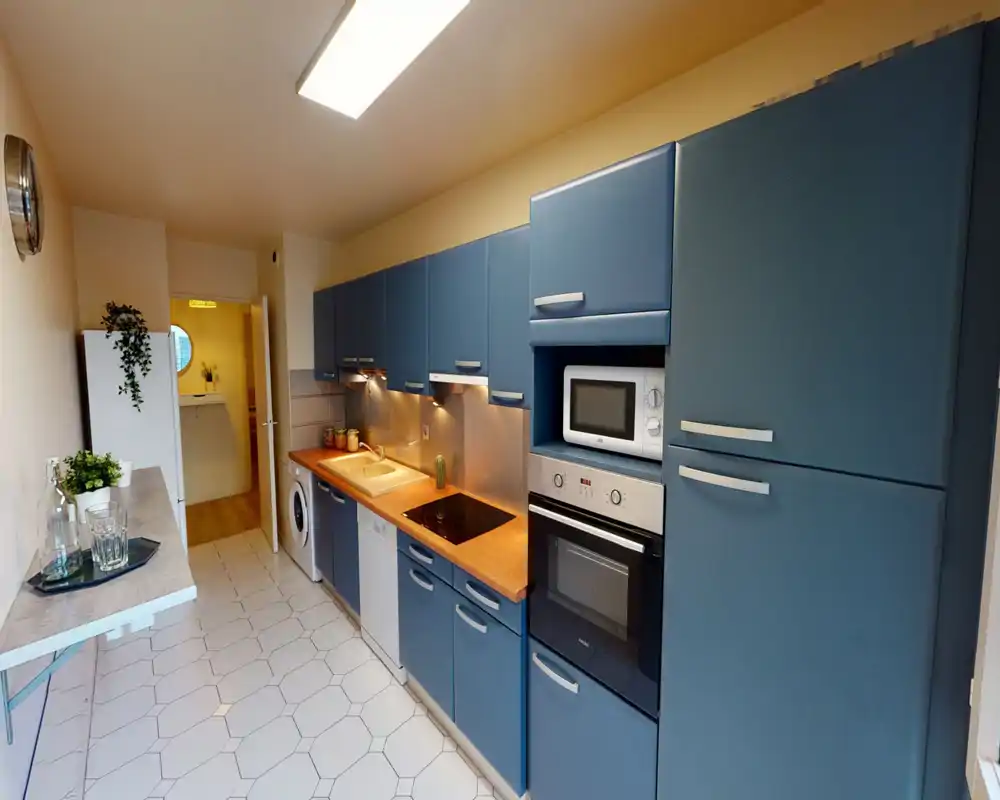 Cucina a corridoio luminosa e moderna con mobili blu, piano in legno, forno e microonde integrati, lavastoviglie e lavatrice; styling curato con illuminazione sotto i pensili e piccole piante.