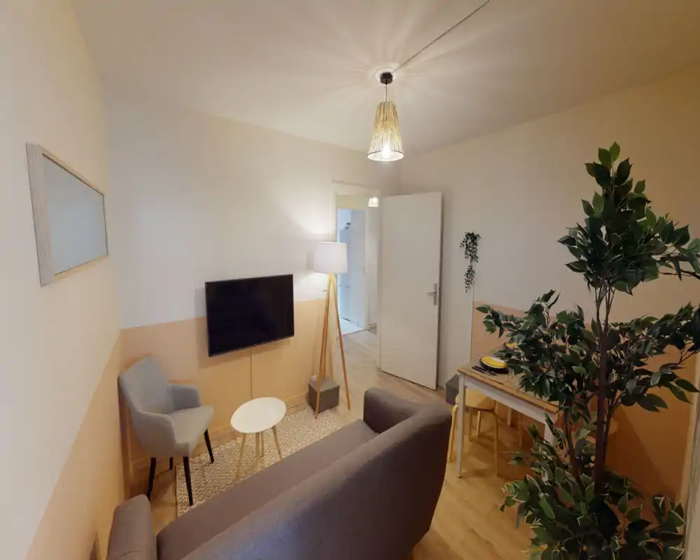 Soggiorno accogliente e ben illuminato con divano, poltrona, TV a parete e un piccolo angolo pranzo. Toni caldi e decorazioni curate creano un’atmosfera confortevole.