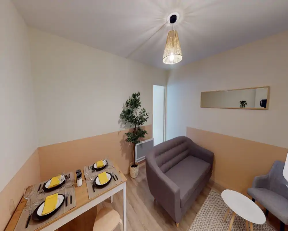 Angolo living compatto e ben arredato con piccolo divano, poltrona e tavolo da pranzo apparecchiato per quattro. Pareti bicolore calde, tappeto e pianta in vaso creano un ambiente accogliente, perfetto per affitti brevi.