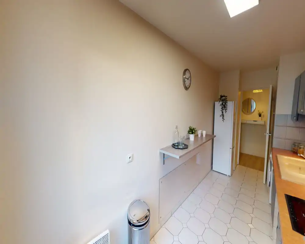 Cucina compatta e luminosa con pavimento piastrellato bianco, piano di lavoro con lavello e piano cottura, frigorifero e un ripiano stretto con piccole piante e bicchieri.