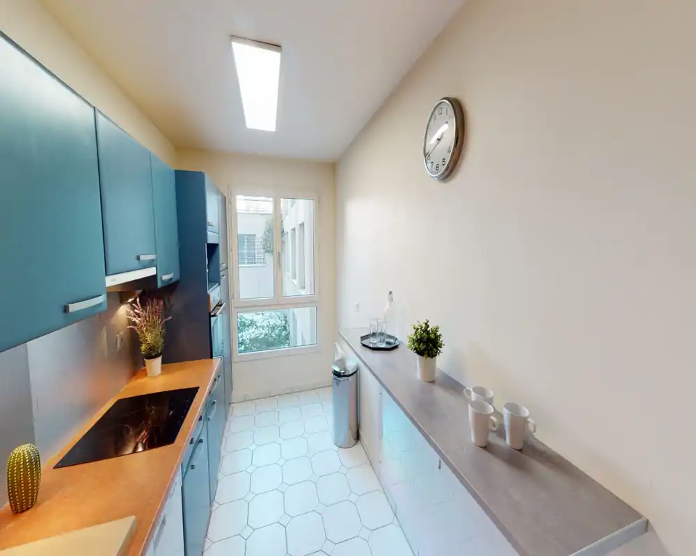 Cucina moderna a corridoio luminosa con pensili turchesi, piano cottura a induzione e lungo bancone con piante e tazze decorative — ben illuminata da luce a soffitto e da una grande finestra.