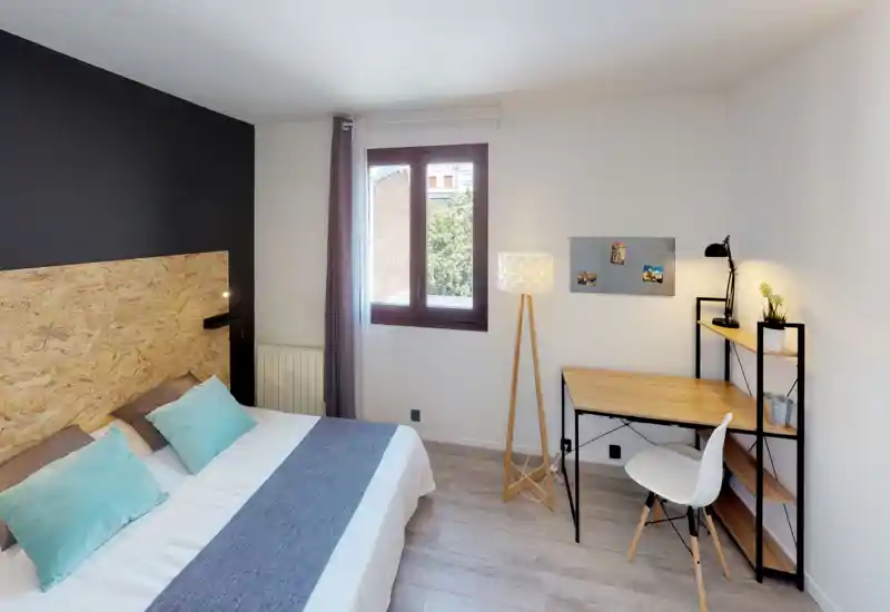 Bright, modern bedroom with a large double bed, textured wooden headboard and a neat workspace by the window — airy natural light and warm lamps create a welcoming stay.