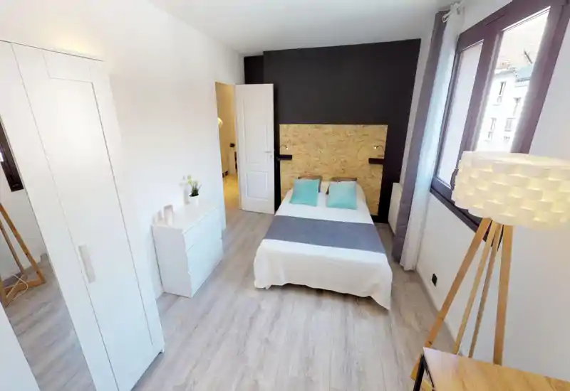 Bright, modern bedroom with a double bed centered against a textured wooden headboard, two aqua pillows, wardrobe and large window providing natural light.