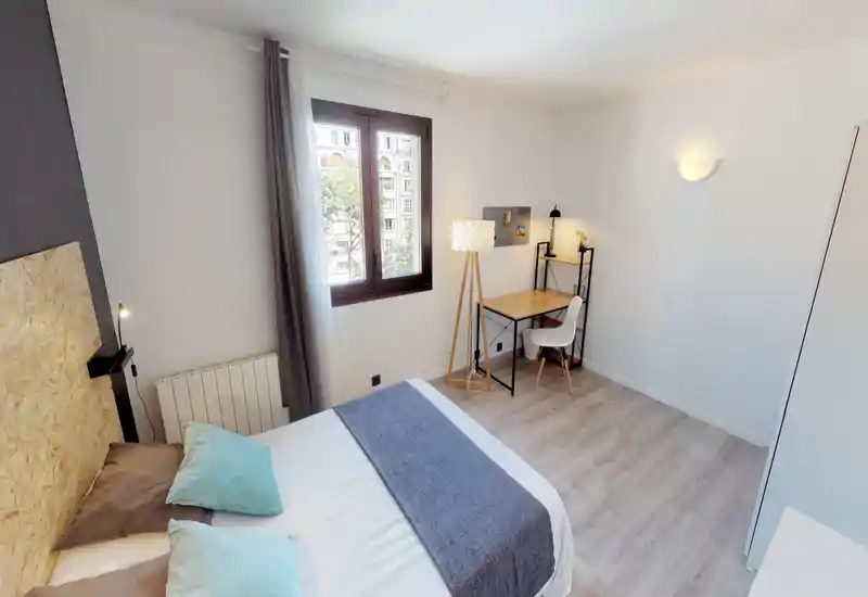 Bright, neatly staged bedroom with a comfortable double bed in the foreground, soft linens and pillows, a window with natural light, and a compact desk corner with lamp — ideal for rest and remote work.