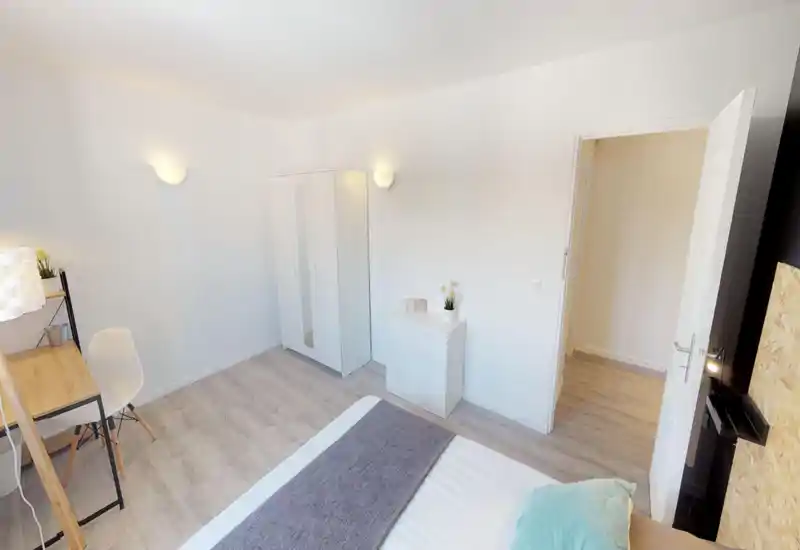 Bright, minimal bedroom with a visible double bed in the foreground, a white wardrobe and dresser, and a small desk. Neutral tones and simple decor create a calm, airy atmosphere.