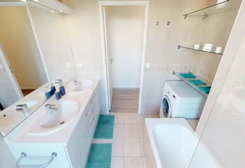 Bright, well-organized bathroom with a double sink vanity, bathtub, and a washing machine tucked under glass shelves. Turquoise towels and mats add a fresh, coordinated accent to the neutral tile finishes.