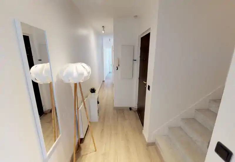 Bright, modern entrance hall with light wood flooring, a standing lamp, wall mirror and a staircase — clean minimalist styling and a long hallway leading further into the home.