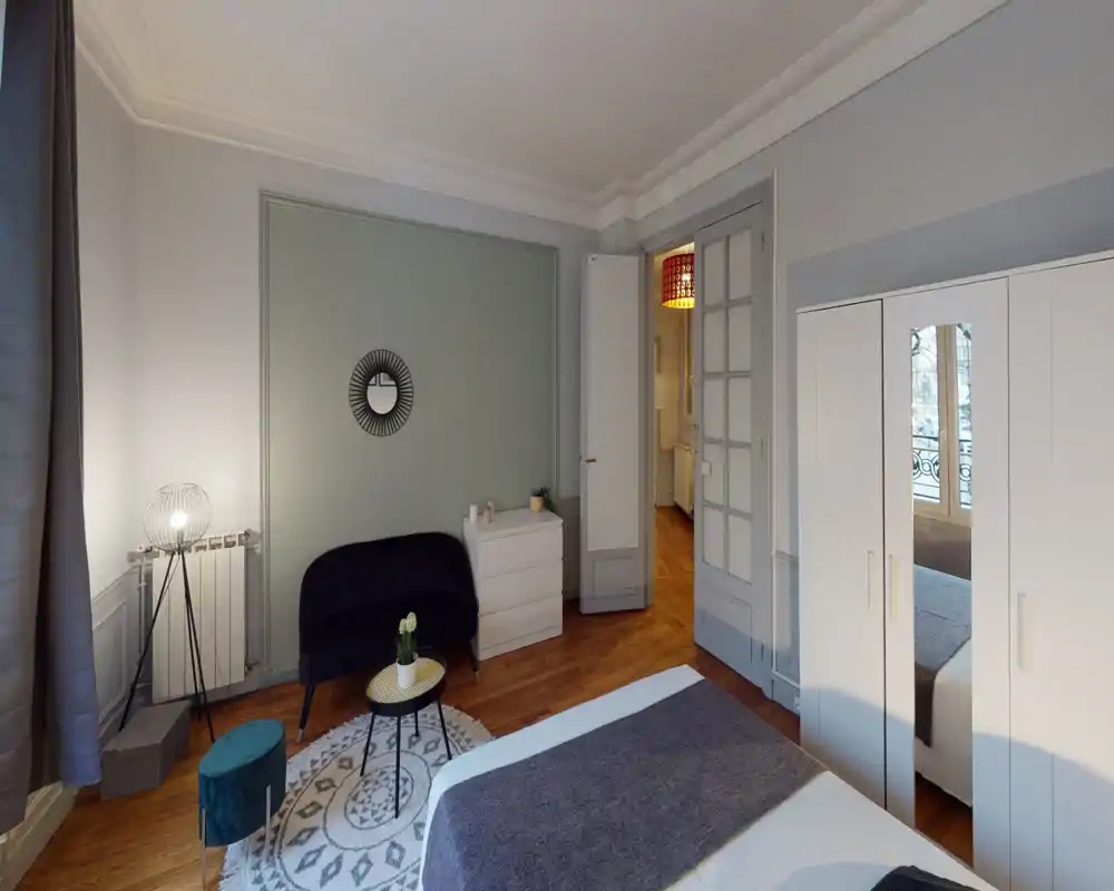 Bright, stylish bedroom with a visible double bed in the foreground, a cozy rug and seating area, and a wardrobe with mirrored door reflecting natural light.