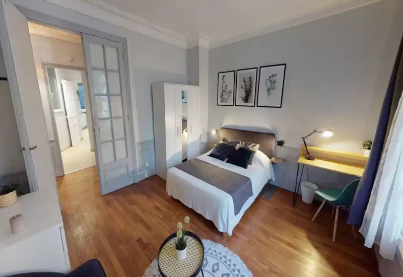 Bright, well-styled bedroom with a double bed, bedside lamps, wardrobe and a small desk by the window. Warm wooden floor and framed botanical prints give a cozy, inviting atmosphere.