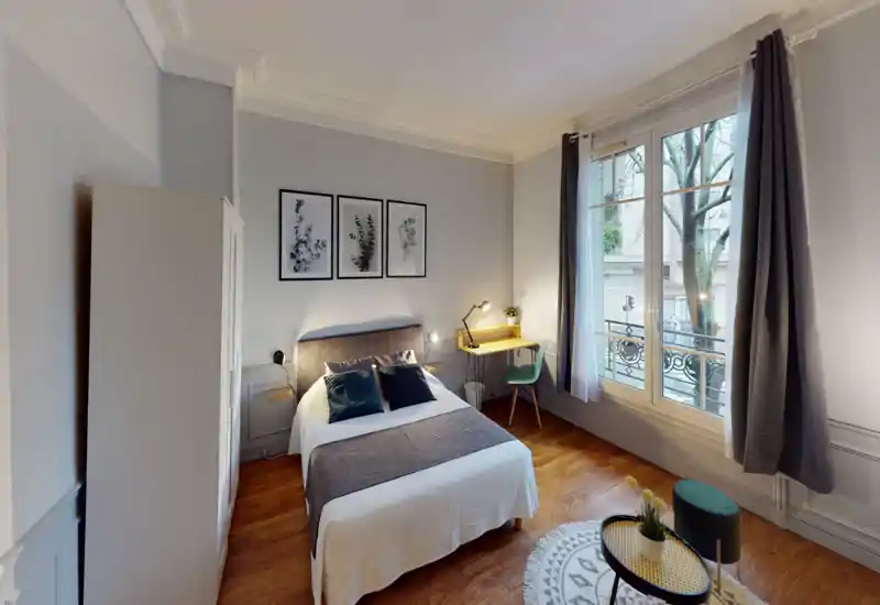 Bright, well-styled bedroom with a central double bed, decorative pillows and framed botanical prints above the headboard. Large window provides natural light and a small desk creates a functional corner — ideal for short-term rentals.