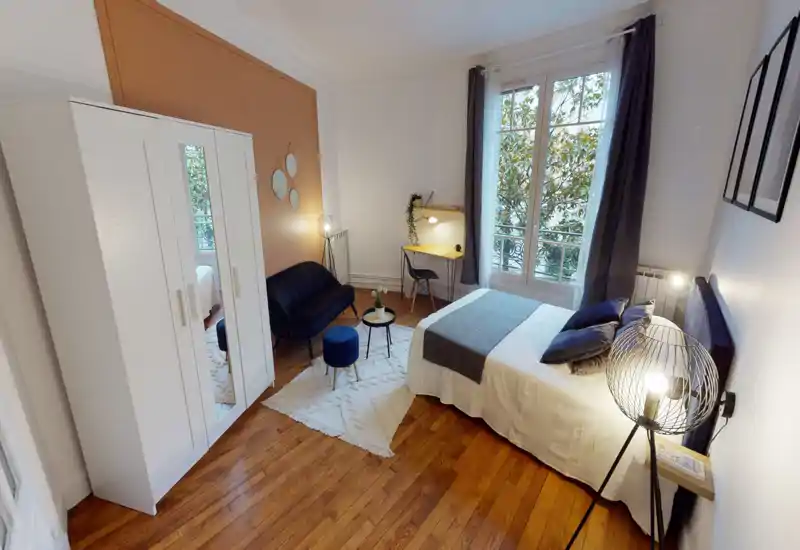 Bright, stylish bedroom with a double bed, soft linens and decorative cushions. The room features a wardrobe with mirror, a small seating area, desk by the window and wooden floors.