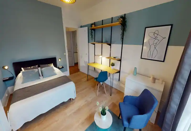 Spacious, well-lit bedroom with a double bed, bedside lamps, a desk with shelving and a comfortable armchair — bright hardwood floors and tasteful blue-accented decor.