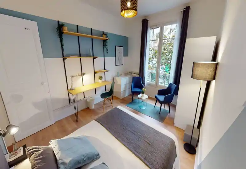 Bright, well-styled bedroom with a neatly made bed in the foreground, a wooden desk and shelving on the left, and a seating area by the window. Natural light and lamps create a warm, inviting atmosphere ideal for listings.
