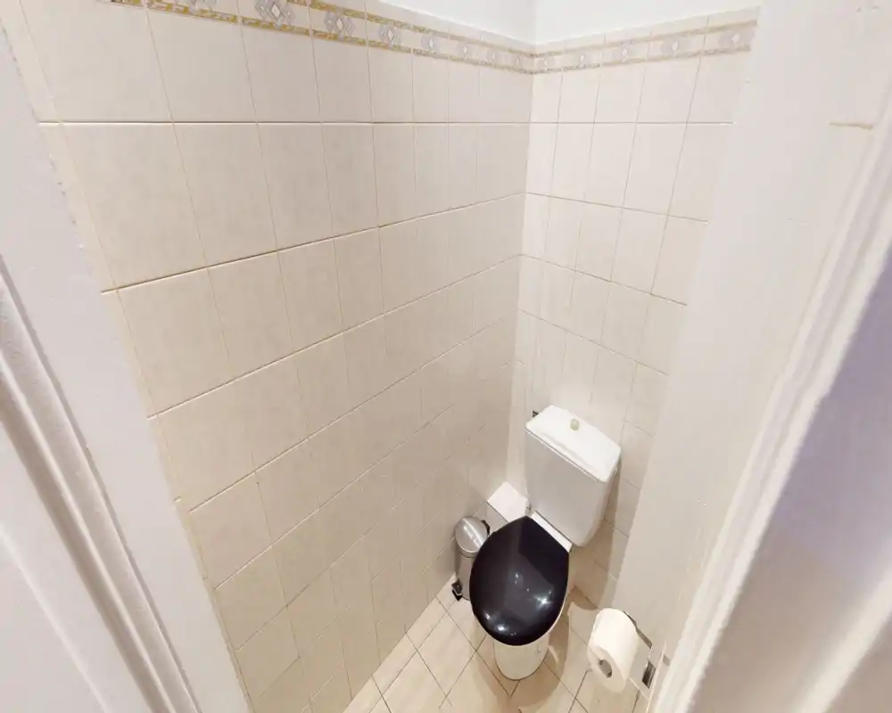 Small tiled bathroom with a compact toilet, black seat, waste bin and toilet paper holder; bright neutral tiles and clean, simple finishes.