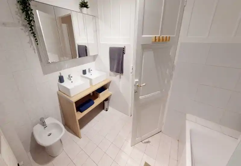 Clean, well-lit bathroom with a double sink vanity, large mirror, bidet and bathtub. Neutral tiles and wooden shelving create a neat, functional space.