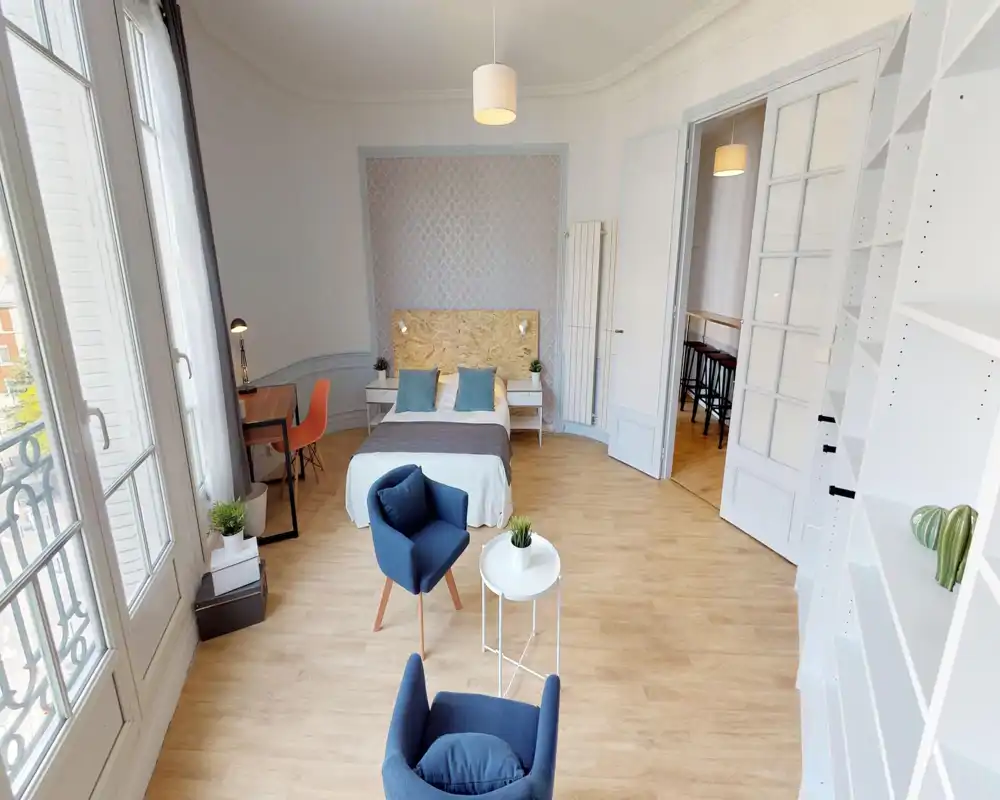 Camera da letto luminosa e di design con letto matrimoniale centrale, comodini e ampie finestre a balcone che garantiscono molta luce naturale. L'ambiente include poltrone moderne, una piccola scrivania e scaffalature integrate per un aspetto arioso e pulito.