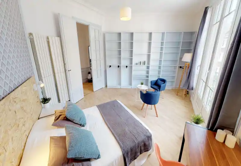 Camera da letto luminosa e di stile con un ampio letto matrimoniale in primo piano, morbidi cuscini e una testiera in legno; è visibile un'area salotto accogliente e scaffalature a parete, con grandi finestre che forniscono molta luce naturale.