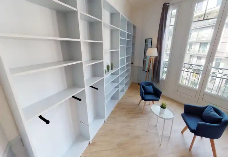 Zona giorno luminosa e moderna con due poltrone blu, un piccolo tavolino bianco e scaffalature integrate; ampie finestre che illuminano l'ambiente.