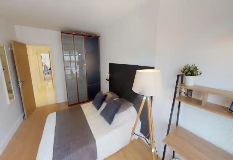 Bright, modern bedroom with a double bed dressed in neutral linens, a tall wardrobe, and a wooden tripod floor lamp — clean, well-staged space ideal for short-term rentals.