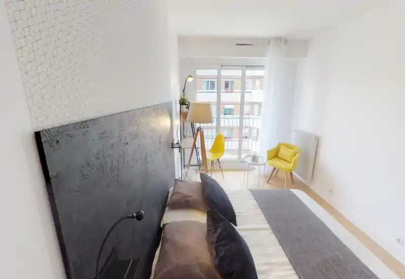 Bright, modern bedroom featuring a large bed with textured dark headboard, layered bedding and a cozy seating corner by a window that fills the room with natural light.
