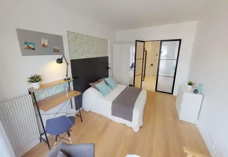 Chambre lumineuse et bien mise en scène avec lit double, coussins décoratifs et espace bureau. Sol en bois et murs clairs pour une atmosphère moderne, idéale pour une annonce locative.