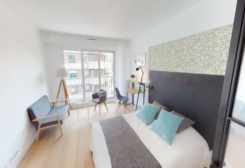 Chambre moderne et lumineuse avec lit double, tête de lit décorative et deux coussins turquoise ; coin salon et bureau près d’une grande fenêtre offrant une belle lumière naturelle.