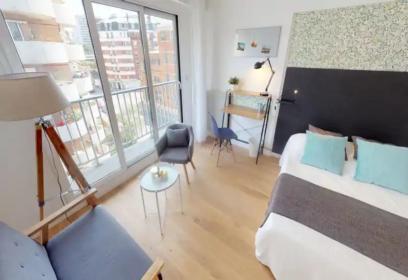 Chambre moderne et lumineuse avec lit double, coussins décoratifs et tête de lit à motif. De grandes portes-fenêtres apportent beaucoup de lumière naturelle; un bureau et un coin détente la rendent pratique.