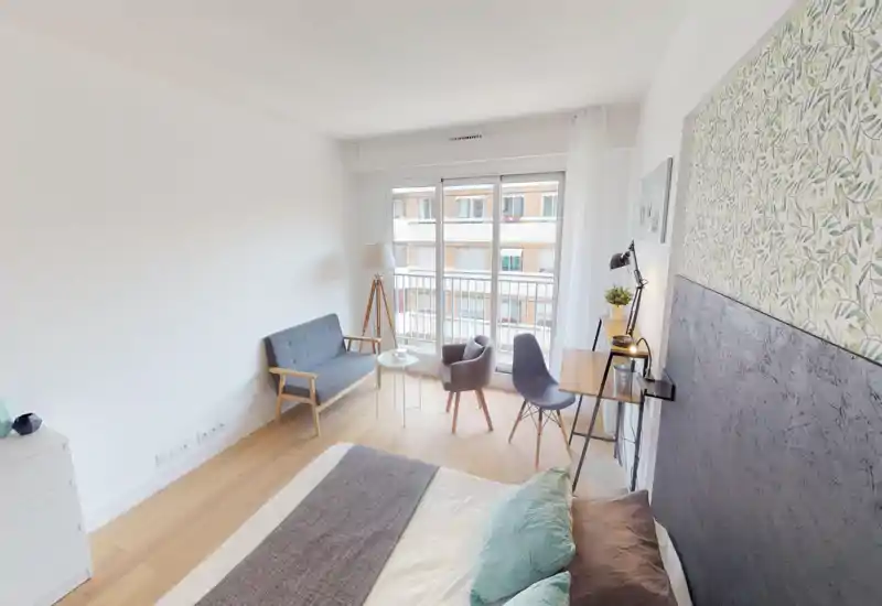 Chambre moderne et lumineuse avec une grande fenêtre et accès au balcon. Elle comprend un lit double confortable avec coussins, un petit canapé et un coin bureau — idéale pour se reposer ou travailler légèrement.
